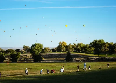 Row of golfers on a driving range looking out towards numerous drifting hot air balloons.