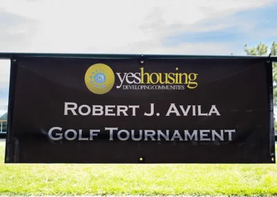 Sign for the Robert J. Avila Golf Tournament with Yes Housing logo overlaid on a yellow golf ball..